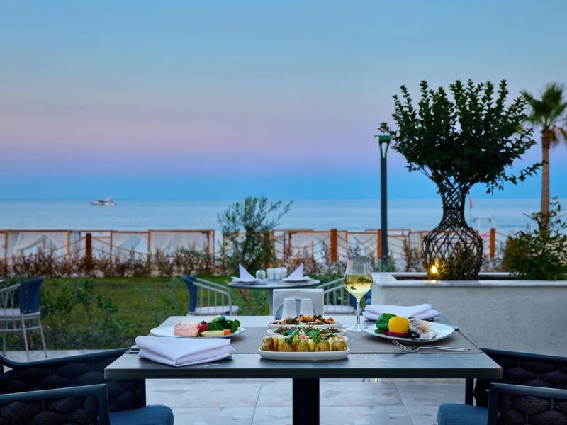 Tisch mit Essen auf Terrasse mit Blick aufs Meer, blauer Himmel und Pflanzen im Hintergrund.