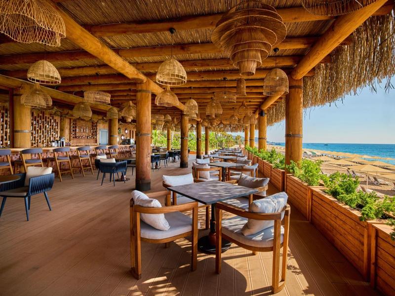 Restaurant mit Holztischen und Korbsesseln unter einem Dach aus Palmblättern am Meer.