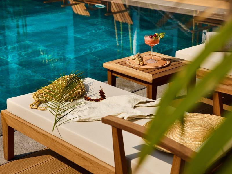 Schöne Holzliege mit Polster am Poolrand, darauf liegen Blumen und ein Cocktail auf dem Beistelltisch.