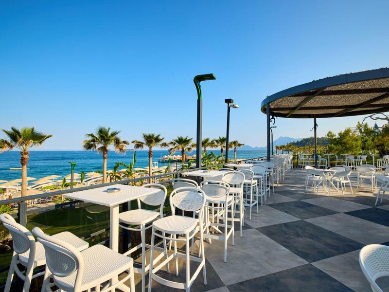Terrassenrestaurant mit weißen Stühlen und Tischen, Palmen und blauem Meer im Hintergrund.