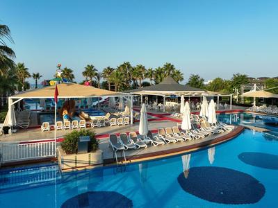Freizeitpool mit vielen Sonnenliegen, weißen Sonnenschirmen und Palmen vor klarem blauem Himmel.