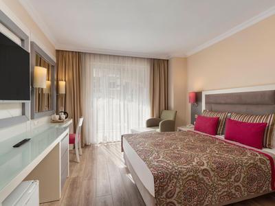 Modern eingerichtetes Hotelzimmer mit Doppelbett, TV, Schreibtisch und Balkon mit Vorhängen.