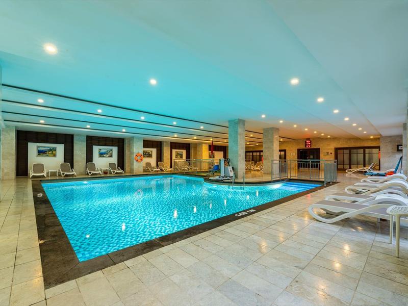 Área moderna con piscina cubierta en hotel, con tumbonas y luz brillante.