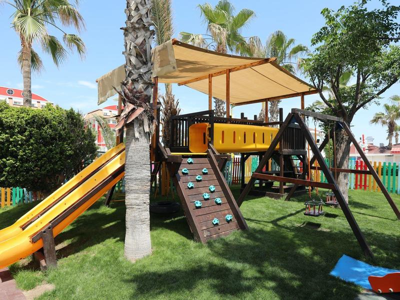 Área de juegos infantiles con tobogán, muro de escalada y columpios en un jardín con palmeras.