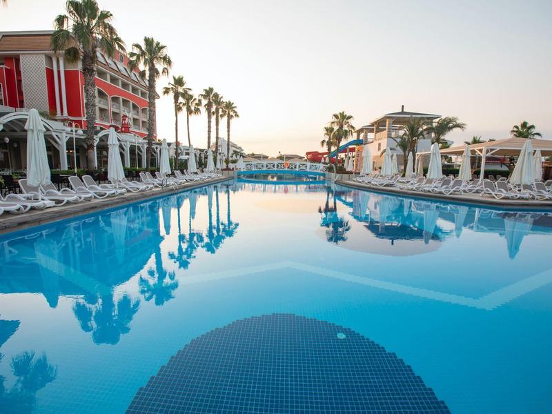 Gran piscina exterior de un resort con palmeras y tumbonas al atardecer.