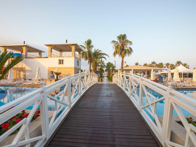 Puente de madera sobre la piscina en un resort soleado con palmeras y edificios blancos.