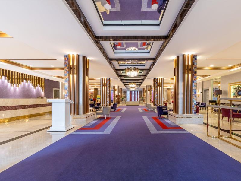 Un amplio pasillo luminoso del vestíbulo del hotel con alfombras azules, columnas doradas y muebles modernos.