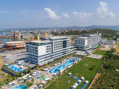 Grand bâtiment d'hôtel avec piscine et parasols dans un environnement verdoyant sous un ciel clair.