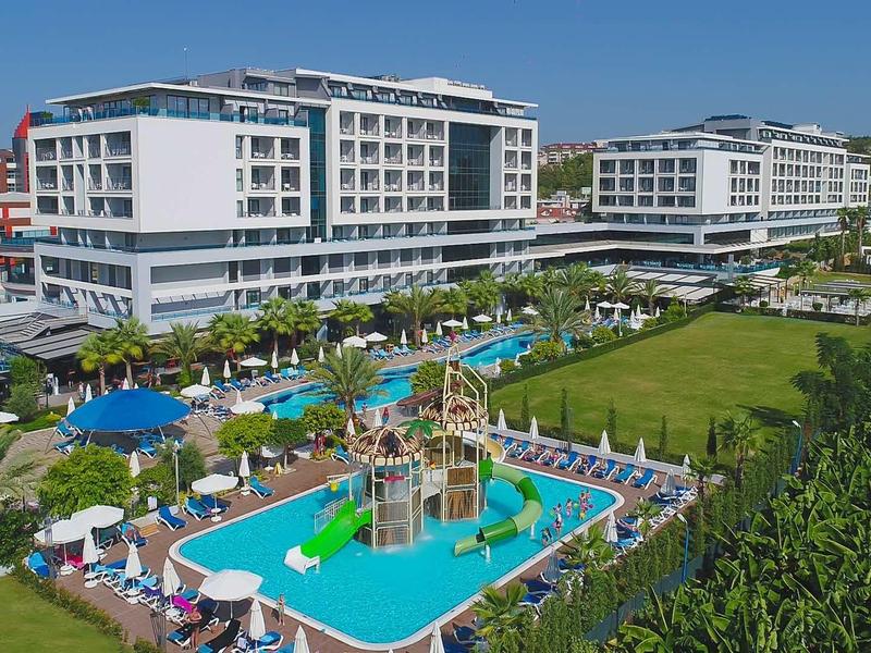 Complexe hôtelier moderne avec piscine extérieure, aire de jeux pour enfants et transats sous un ciel bleu.