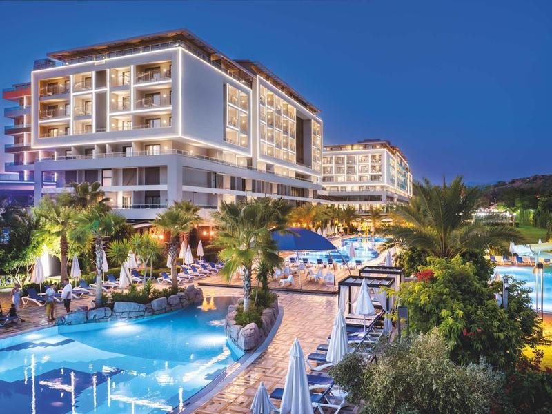 Grand complexe hôtelier avec piscines éclairées et plantes tropicales au coucher du soleil.