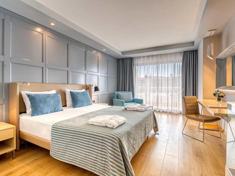 Chambre d'hôtel moderne avec lit double, fauteuils, bureau et lumière naturelle provenant de grandes fenêtres.