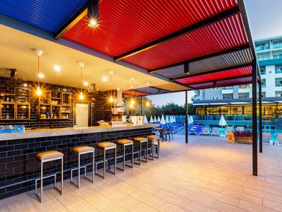 Moderne Bar mit Hockern, rotem Sonnendach und Poolbereich in einem Stadthotel bei Abendlicht.