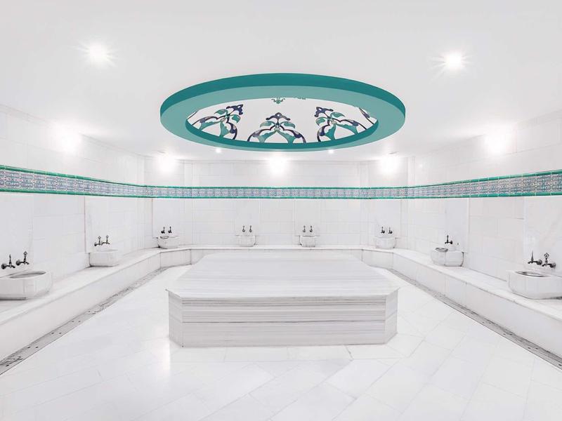 Großer weißer Hammam mit Marmor-Ottoman in der Mitte und türkisfarbener Deckenverzierung.