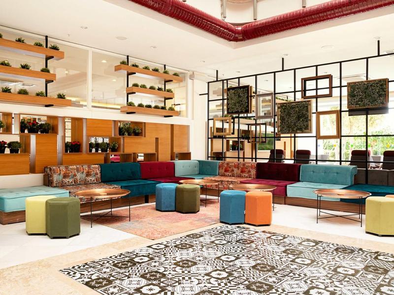 Großer, heller Raum mit bunten Sitzwürfeln, Sofas, Teppichen und vielen Pflanzen an den Wänden.