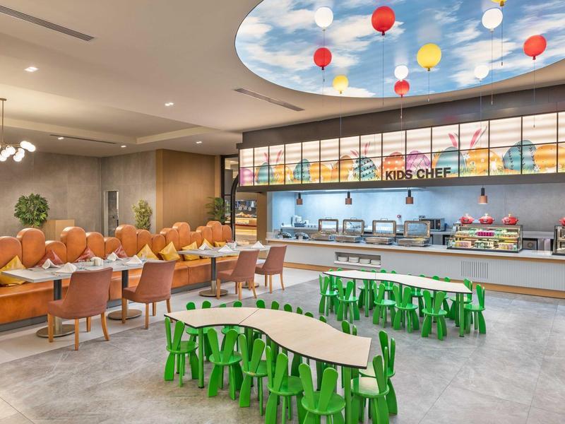 Sala de desayuno moderna con lámparas coloridas, mesas para niños y asientos cómodos en tonos cálidos.