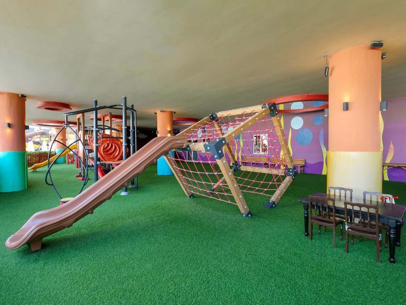 Área de juegos cubierta colorida con toboganes, redes para escalar y zona de asientos sobre alfombra verde.