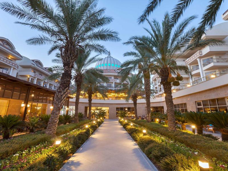 Camino iluminado entre palmeras conduce a un moderno edificio hotelero al atardecer.
