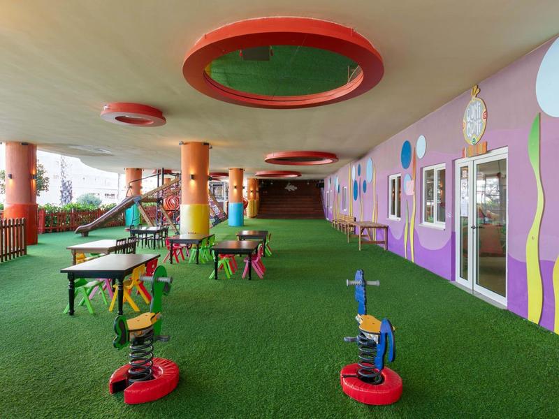 Área de juegos cubierta con estructura colorida y césped artificial verde en entorno hotelero.