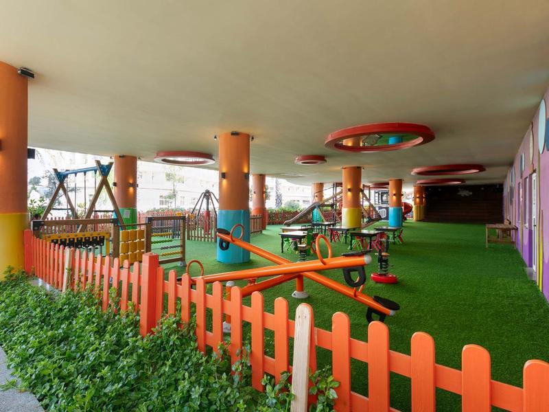 Área de juegos cubierta con césped artificial, columpios y cerca naranja en un hotel.