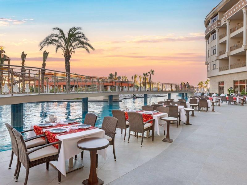 Restaurante al aire libre con vista a la piscina y palmeras al atardecer en un resort hotelero.