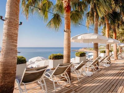 Sonnige Terrasse mit Liegestühlen, weißen Sonnenschirmen und Palmen an einem ruhigen Meer.