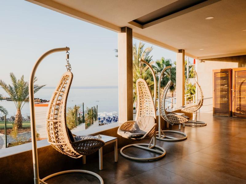 Sunlit balcony with hanging rattan chairs overlooking palm trees and sea at resort.