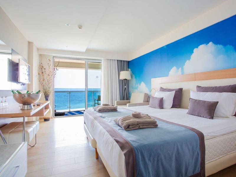 Bright hotel room with twin beds, ocean view balcony, and blue sky mural on the wall.