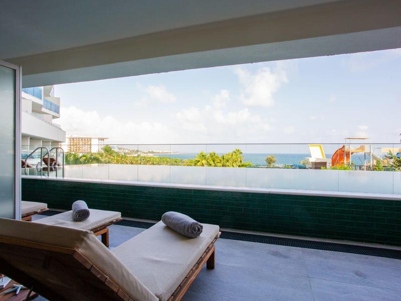Balcony with lounge chairs overlooking a pool and ocean under a blue sky.