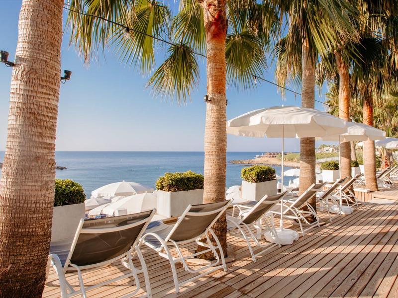 Ocean view with sun loungers and umbrellas on a wooden deck under palm trees.