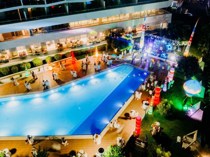 Illuminated outdoor pool with guests in front of a multi-story hotel building at night.