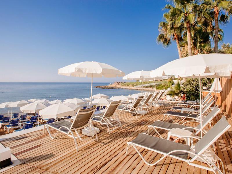 Empty lounge chairs and white umbrellas on wooden deck overlooking calm sea under clear sky.