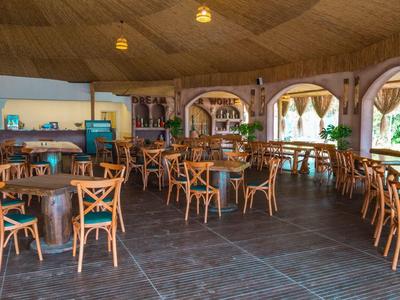 Restaurace s dřevěnými stoly a židlemi pod dřevěnou střechou, světlý interiér s otevřenými okny.