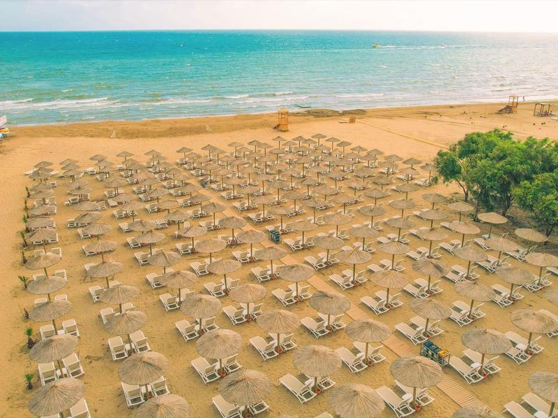 Pláž s řadami lehátek a slunečníků u moře a zalesněné oblasti.