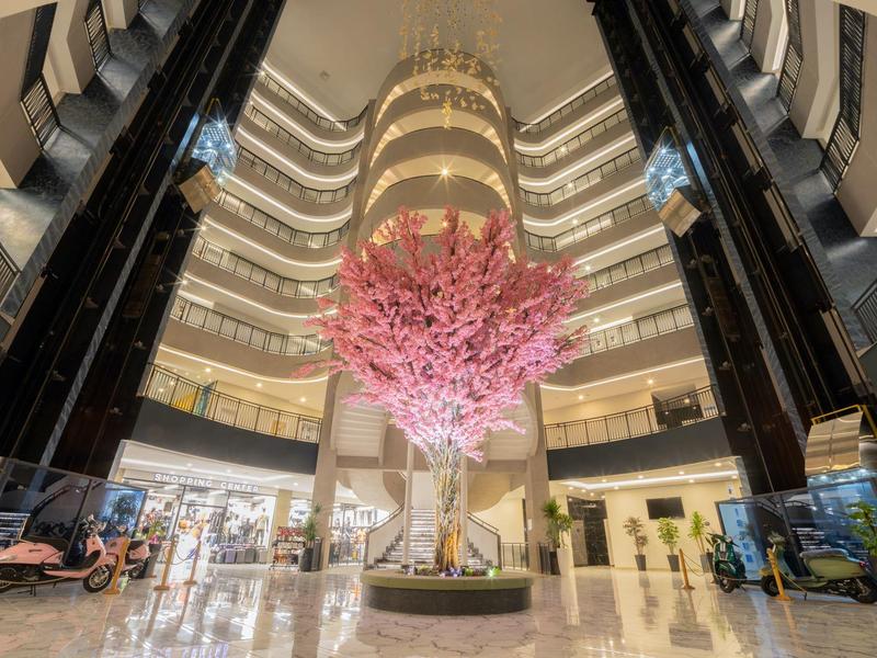 Interiér moderního hotelu s velkým růžovým stromem uprostřed a panoramatickými výtahy.