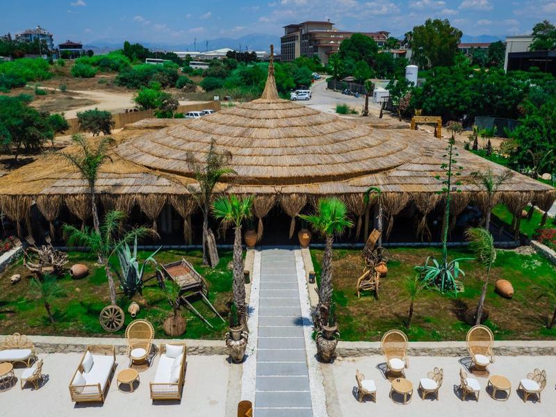 Letecký pohled na bazén obklopený lehátky a venkovní restaurací s listovou střechou.