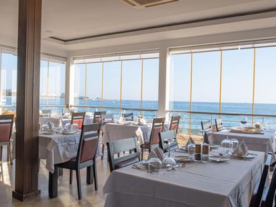 Světelná restaurace se skleněnými okny a výhledem na moře, prostřené stoly s bílými ubrusy.