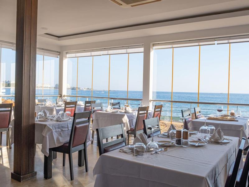 Světelná restaurace se skleněnými okny a výhledem na moře, prostřené stoly s bílými ubrusy.