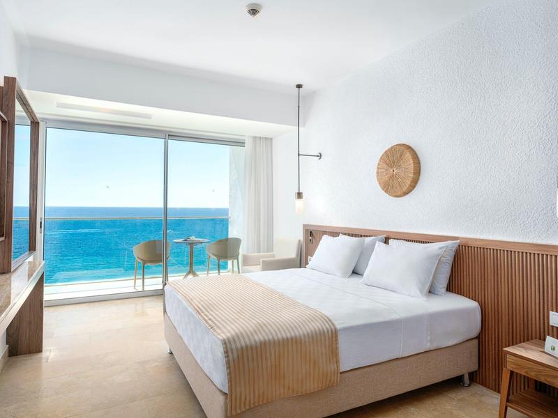 Světle hotelový pokoj s manželskou postelí, balkonem a výhledem na moře.