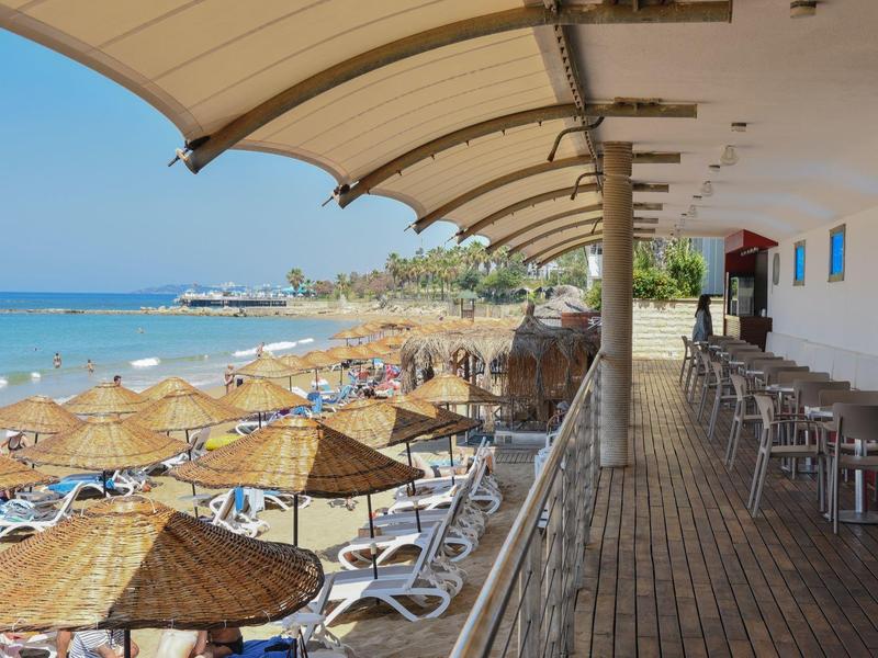 Zastřešená terasa s jídelními stoly na pláži s výhledem na moře a lehátka se střešními krytinami z trávy.