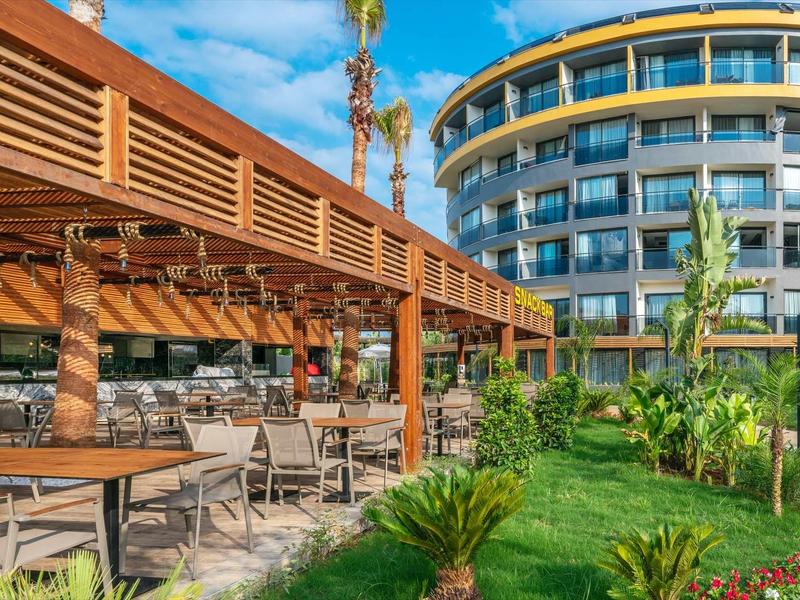 Outdoor hotel terrace with tables, chairs, pergola, and modern curved building in background.