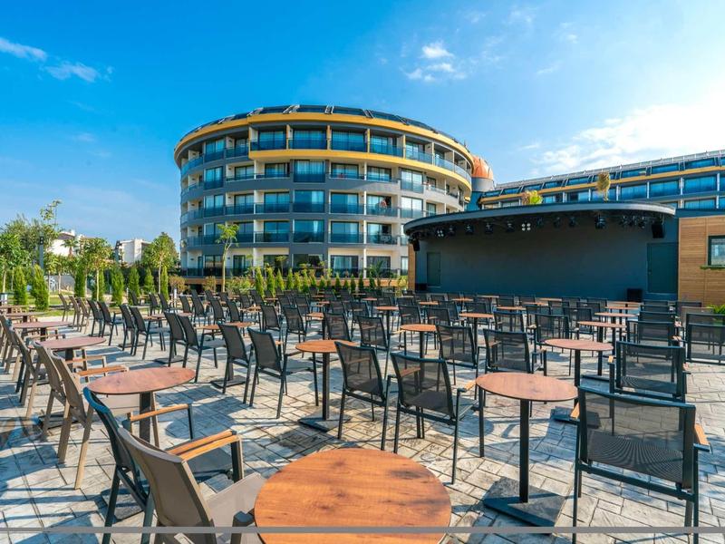 Outdoor seating area with tables and chairs in front of a modern circular hotel building under blue sky.