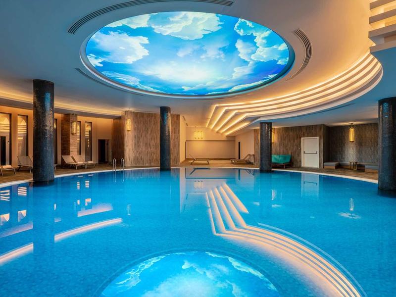 Indoor pool with a sky-themed ceiling light and elegant lighting in a hotel spa area.