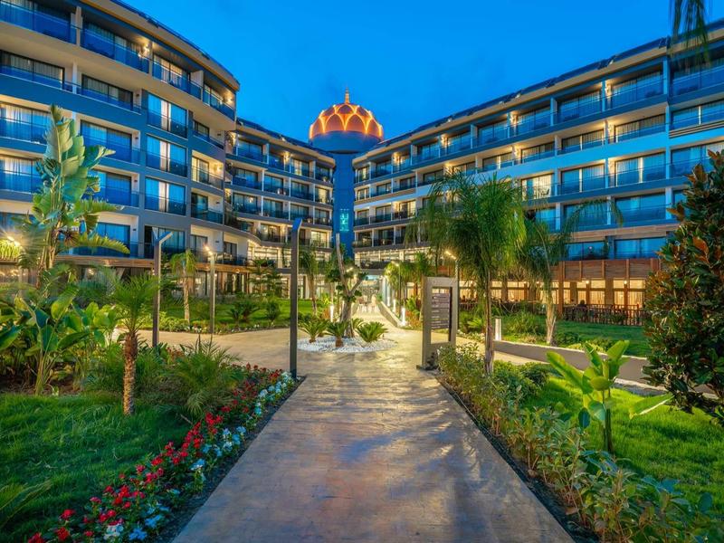 Modern hotel facade with glass balconies, lit at dusk, surrounded by lush greenery and pathway.
