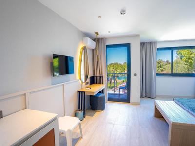 Lichte hotelkamer met bureau, stoel, tv en balkondeur met uitzicht op bomen.