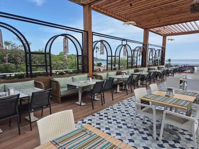Overdekt buitenterras met tafels en stoelen, uitzicht op zee en blauwe lucht.
