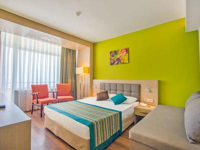 Lichte hotelkamer met groene accentmuur, tweepersoonsbed, oranje stoelen en groot raam met doorschijnende gordijnen.