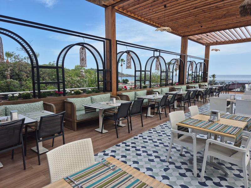 Overdekt buitenterras met tafels en stoelen, uitzicht op zee en blauwe lucht.