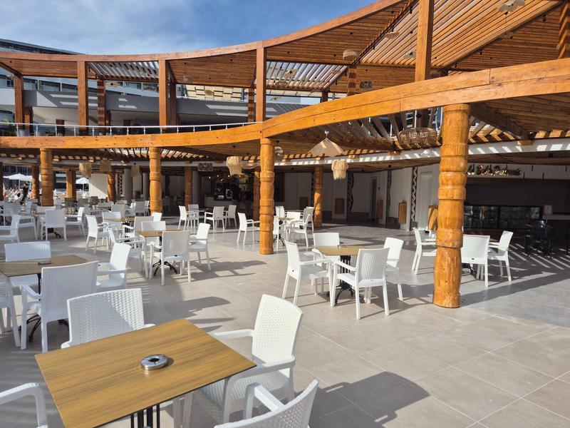 Open eetgedeelte met witte stoelen en houten tafels onder een houten pergola dak.