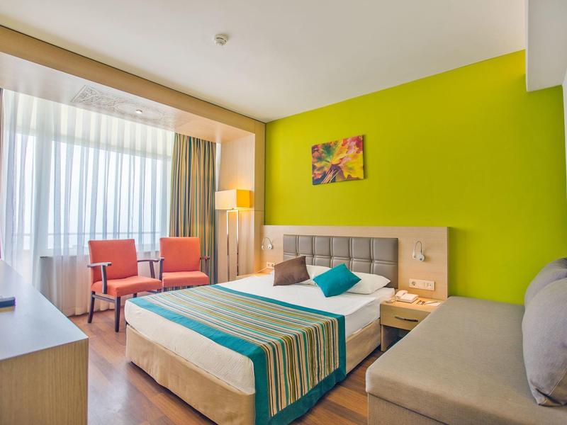 Lichte hotelkamer met groene accentmuur, tweepersoonsbed, oranje stoelen en groot raam met doorschijnende gordijnen.