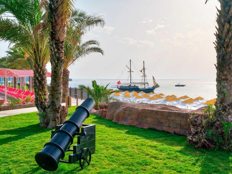 Výhled z zeleného trávníku s dělem mezi palmami na slunnou pláž a pirátskou loď na vodě.